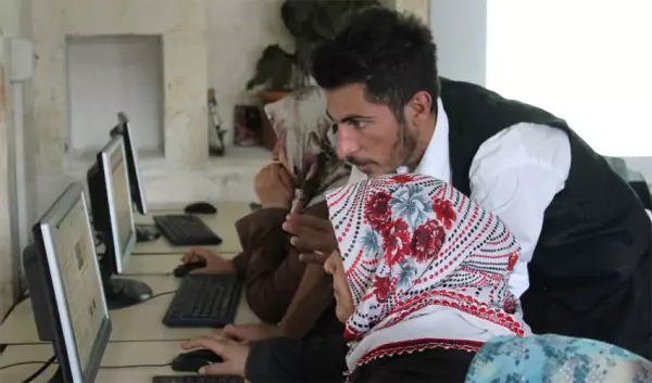 Bilgisayarın önünde oturan bir kadın ve ayakta ekrana bakan bir erkek bulunuyor.